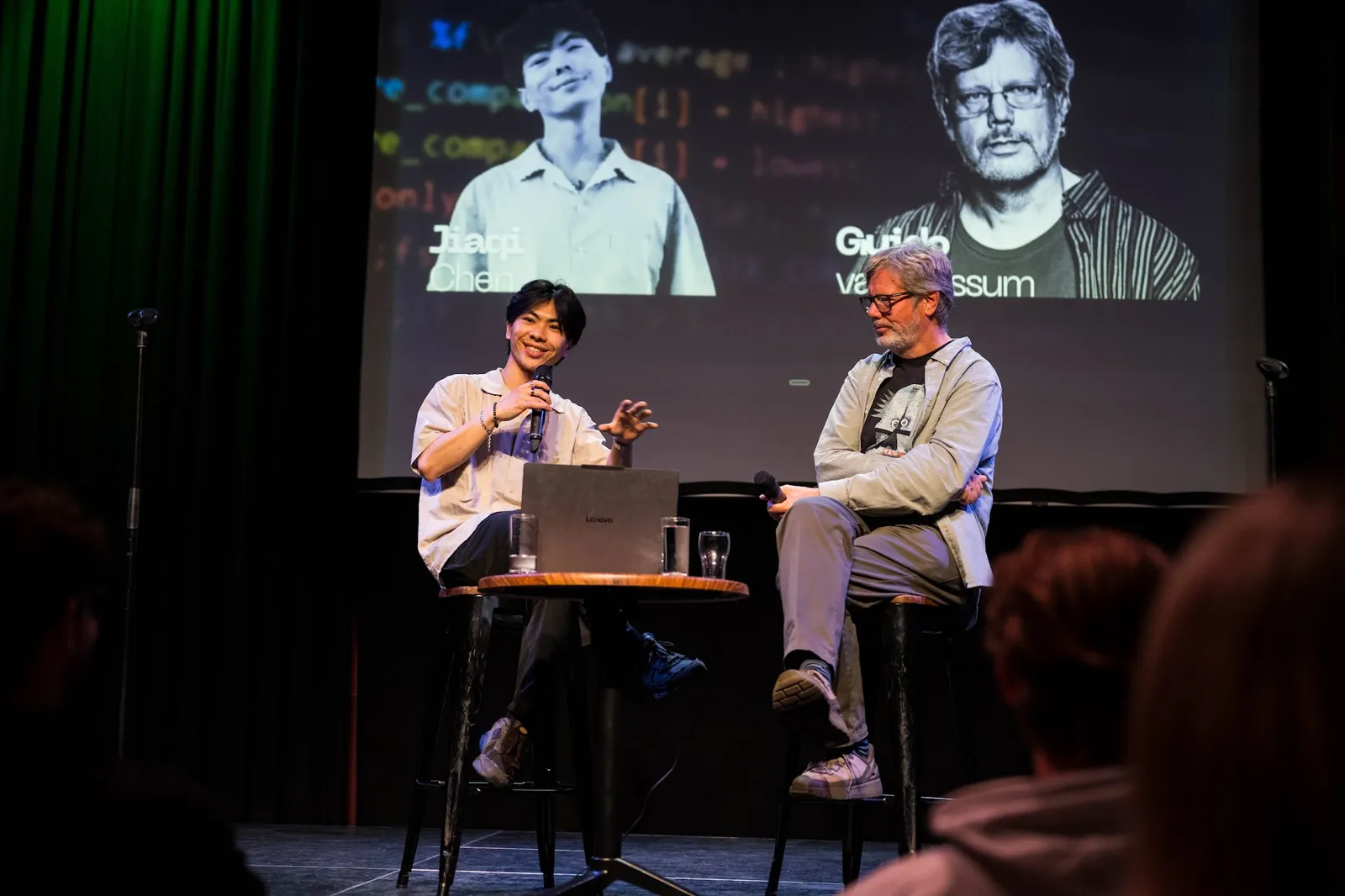 Jiaqi Chen interviewing Guido van Rossum on stage, both holding microphones, with their names projected behind them